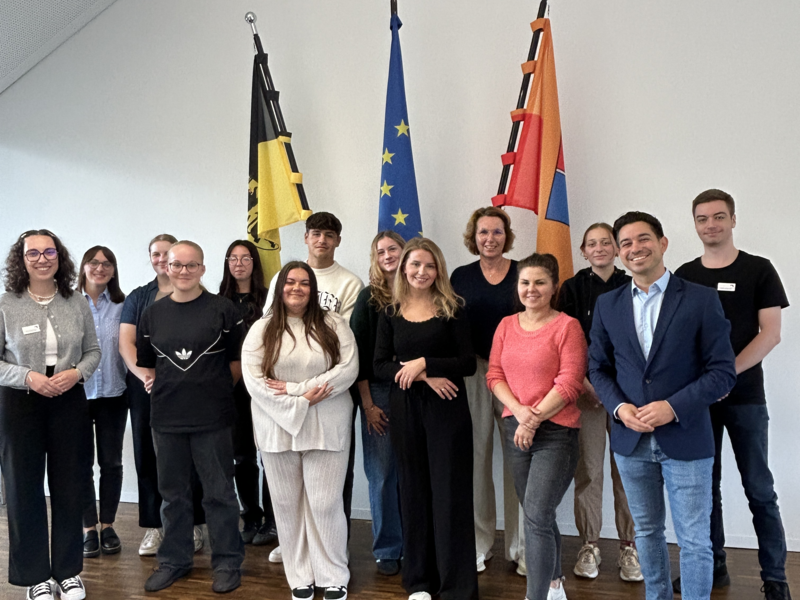 Eine Gruppe von Personen posiert lächelnd für ein Foto in einem Raum mit Holzboden und mehreren Flaggen, darunter die der Europäischen Union, im Hintergrund.