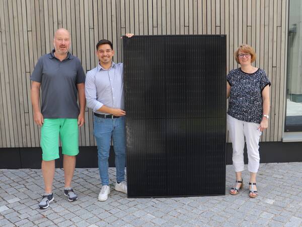 3 Personen mit Photovoltaikmodul vor Gebäude