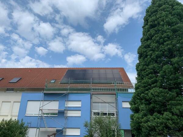 Photovoltaikanlage auf Satteldach auf mehrstöckigem Gebäude mit Baum rechts und wolkigem Himmel