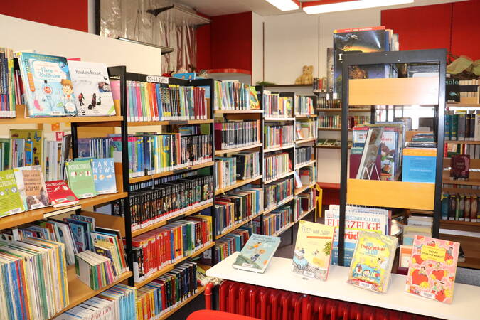 Gang in Bücherei Wössingen, schräg von vorne links nach hinten rechts, im Vordergrund und im Hintergrund viele Bücherregale, ganz vorne ein roter Heizkörper mit einer Art Fensterbrett, auf dem Bücher präsentiert sind