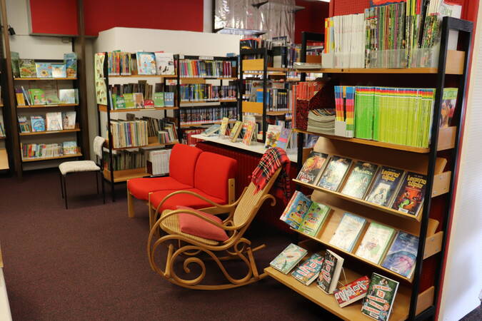 Gang in Bücherei Wössingen, im Vordergrund ein Bücherregal, daneben ein alter Schaukelstuhl und 2 Stühle mit roter Sitzfläche und Lehne. Im Hintergrund weitere Bücherregale