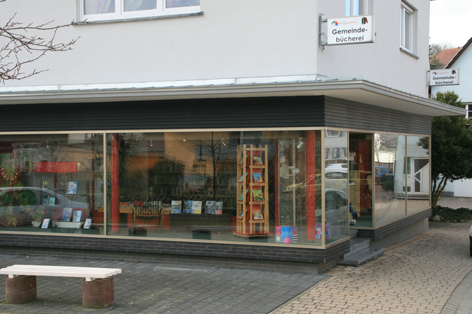 Außenansicht Bücherei Wössingen, durch große Schaufenster sieht man die Bücherregale, im Vordergrund steht vor dem Gebäude eine Sitzbank. Am Gebäude ist das Hinweisschild für die Bücherei zu sehen.