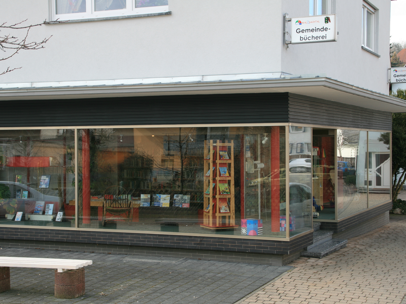 Außenansicht Bücherei Wössingen, durch große Schaufenster sieht man die Bücherregale, im Vordergrund steht vor dem Gebäude eine Sitzbank. Am Gebäude ist das Hinweisschild für die Bücherei zu sehen.