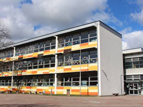 Schulgebäude mit Flachdach und gelb-oranger Fassade, im Vordergrund das Hofpflaster des Schulhofs, am Himmel wolkig mit blauen Lücken.