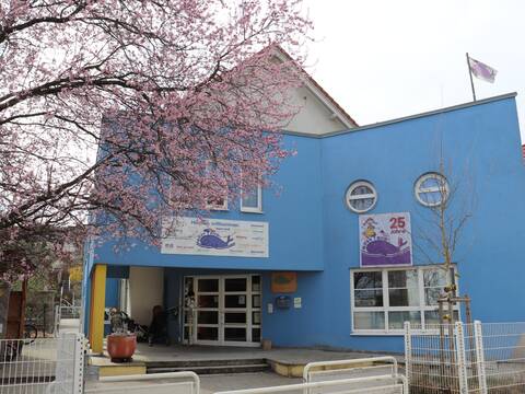 Ein blau farbiges Gebäude, vor diesem ist ein weißer Zaun und nebendran ein Baum mit Kirschblüten.