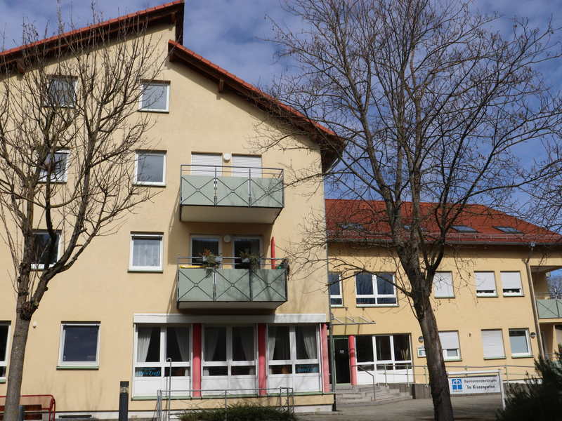 Ein helles großes Gebäude mit vielen Fenster und Balkone, davor sind zwei Bäume.