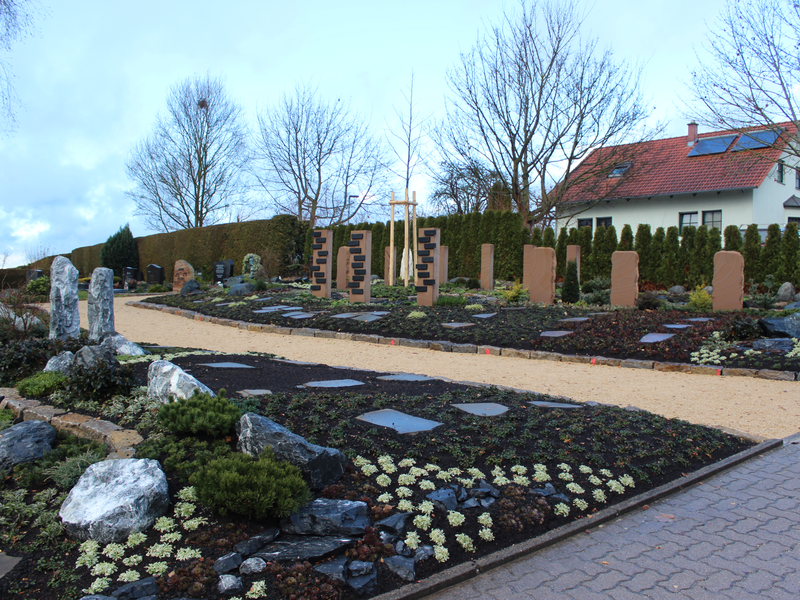 Das Bild zeigt einen sorgfältig angelegten Friedhof mit Grabsteinen und gepflegten Pflanzen inmitten eines ländlichen Bereichs mit einem Haus im Hintergrund.