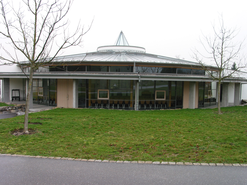 Aussegnungshalle Neuer Friedhof Jöhlingen, im Vordergrund ein Schotterweg, dann Rasenfläche. Dahinter ein moderner, halbrunder Bau mit Glasflächen und Metalldach