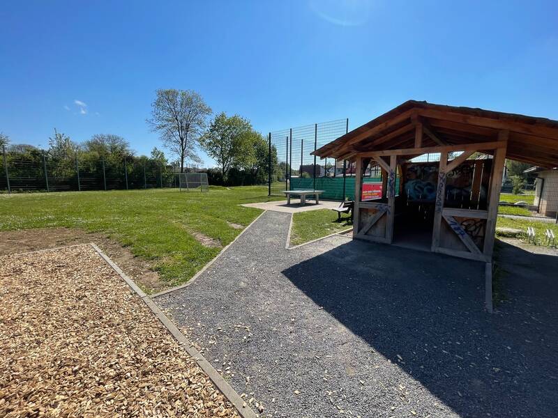 Foto vom Spielplatz an der Wiesenmühle in Jöhlingen. Eine Tischtennisplatte, Holzhütte und grüne Rasenfläche unter blauem Himmel sind zu sehen.