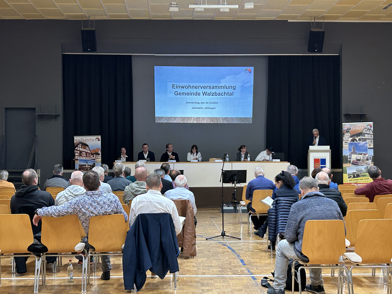 Menschen sitzen in einem Saal bei einer Einwohnerversammlung der Gemeinde Walzbachtal, während vorne eine Präsentation gezeigt wird.