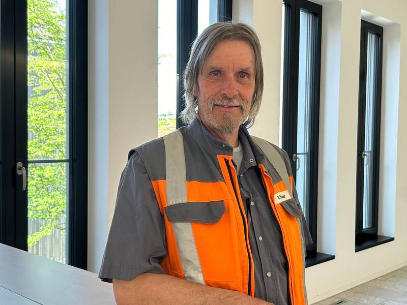 Portrait von Rüdiger Fritscher in orangfarbener Arbeitsjacke, angelehnt an Theke