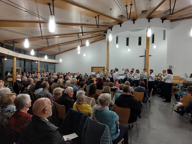 Eine große Gruppe von Menschen sitzt in einem modernen Saal der Aussegnungshalle, während ein Orchester im Bildhintergrund spielt.