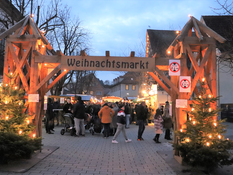 Holzportal mit Schild Walzbachtaler Weihnachtsmarkt. Blick auf den Weihnachtsmarkt auf dem Kirchplatz Jöhlingen, zwischen Kirche und Speyerer Hof. Portal beleuchtet durch zwei Weihnachtsbäume an den Säulen. Im Hintergrund Menschengruppen und die Hütten des Markts