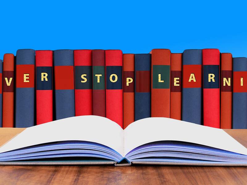 Bücher in roten und blauen Einbänden. Auf den Bücherrücken ist der Satz Never stop learning zu lesen. Ein aufgeklapptes Buch liegt vor den Büchern auf einem Tisch.
