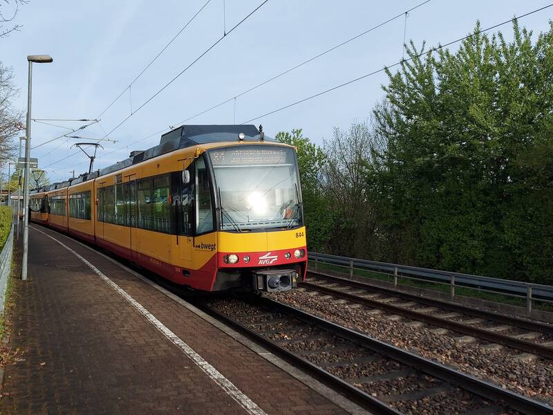 gelb-rote Stadtbahn der Linie S4 wartet an der Haltestelle. Links und Rechts der Gleise ist grüner Bewuchs. Blauer Himmel und Sonnenschein.
