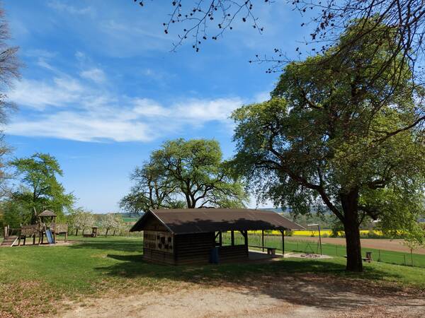 Holzhütte mit Grillstelle zwischen drei großen Bäumen. Im Hintergrund ein Spielgerät für Kinder. Blauer Himmel mit leichter Bewölkung. 