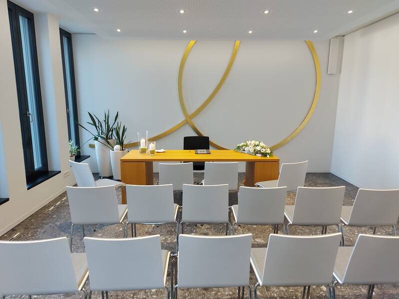 Blick mittig in das Trauzimmer im Rathaus Wössingen. Stühle stehen im Vordergrund. Im Hintergrund der Trausekretär und goldene Ringe an der Wand.