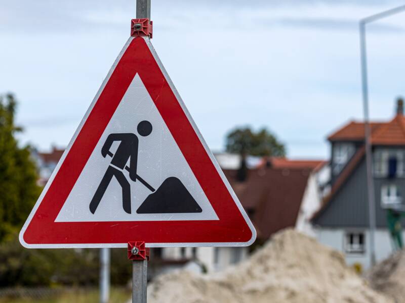 Ein Verkehrsschild, das auf eine Baustelle hinweist. Im Hintergrund befinden sich Sandhäufen auf einer Straße.