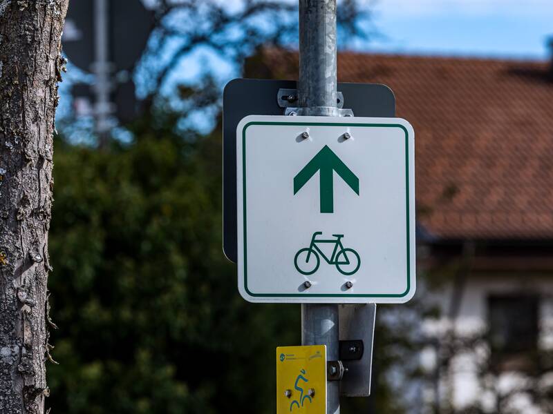 Ein weißes Schild mit einem grünen Fahrrad und Richtungspfeil als Wegweiser für einen Radweg. Das Schild ist an einem Laternenpfosten befestigt.