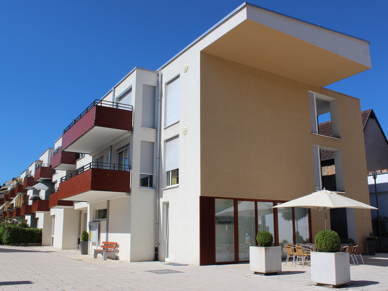 Haus an Gängle Jöhlingen mit weißer Fassage und dunkelroten Balkonen. Davor ein großer Platz mit Schirm und Blumenkübeln