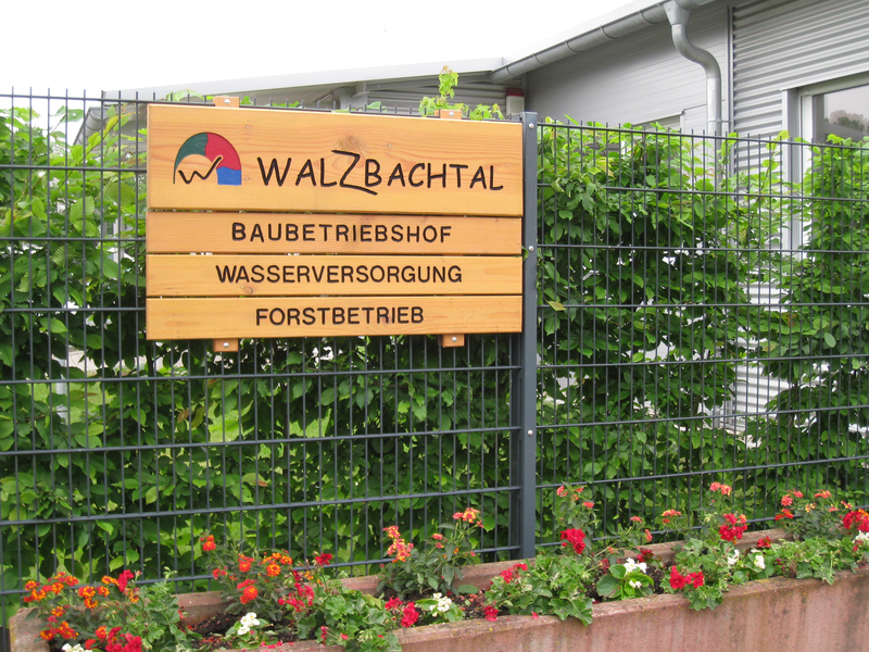 Holzschild mit Walzbachtal Schriftzug und Hinweisschildern. Dahinter Gebüsch, vorne Blumen