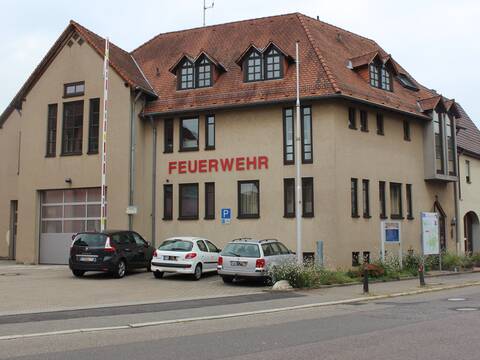 Feuerwehrhaus Jöhlingen mit beige Fassade und Schriftzug. Davor drei Autor und eine geöffnete Schranke