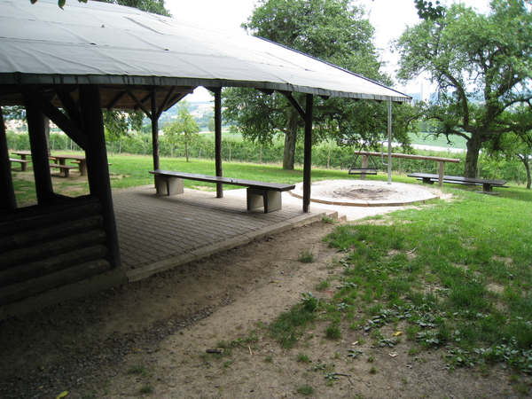 Im Vordergrund ist eine Hozhütte mit einer Holzbank, im Hintergrund befindet sich eine Grillstelle. Drumrum befindet sich eine grüne Wiese und grüne Bäume