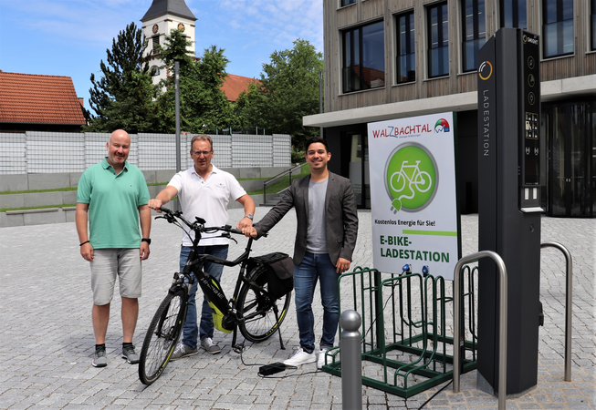 Bürgermeister Özcan und zwei Mitarbeiter der Gemeinde auf dem Radhausplatz mit einem E-Fahrrad davor.  Rechts daneben eine Aufladestation