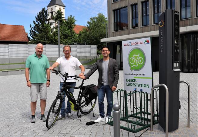 Bürgermeister Özcan und zwei Mitarbeiter der Gemeinde auf dem Radhausplatz mit einem Fahrrad davor.  Rechts daneben eine Aufladestation