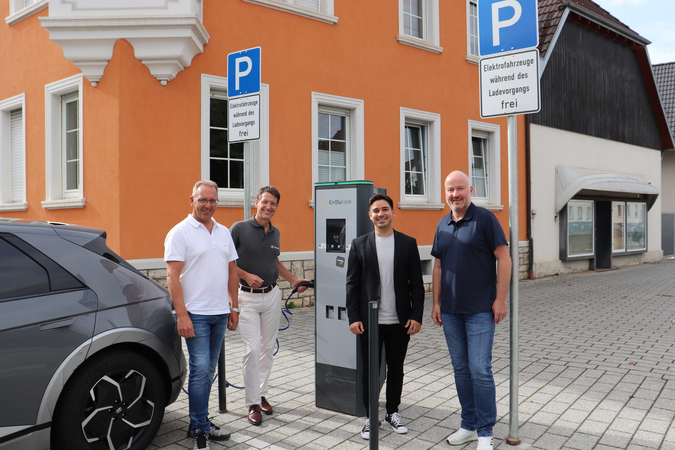 Vier Personen stehen auf dem Kirchplatz. Links davor ein graues Fahrzeug, dass an die E-Ladestation angeschlossen wird