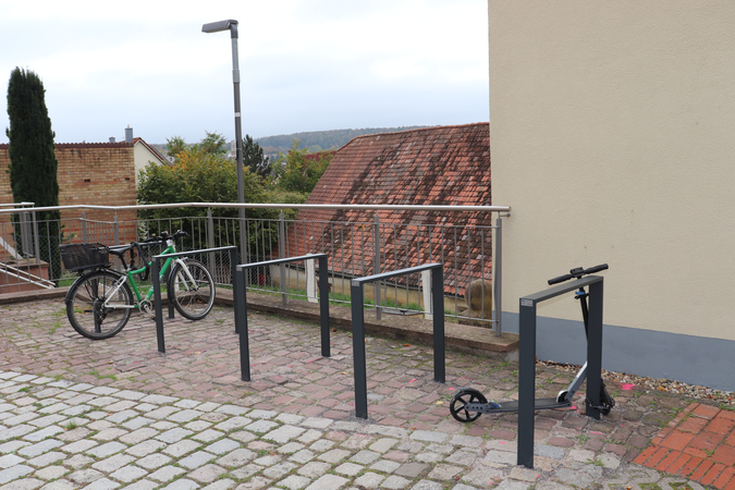 Vier Fahrständer. An einem ist ein Roller angelehnt. Am anderen wurde ein Fahrrad angeschlossen.