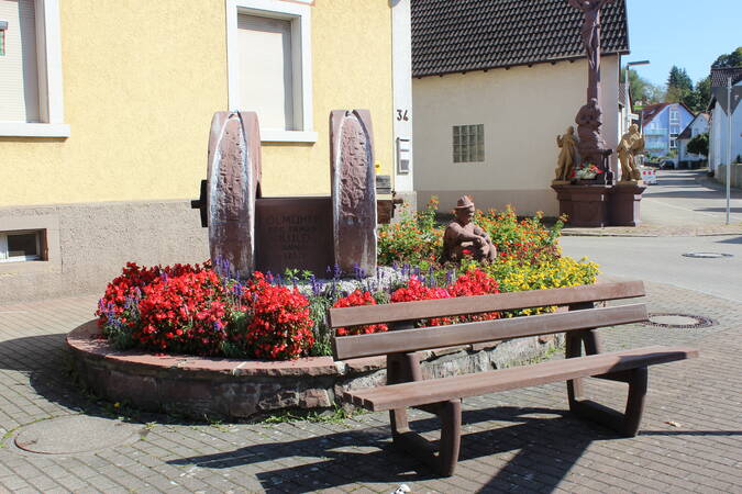 Hirtenbrunnen in Jöhlingen, Im Hintergrund ein Wegekreuz und vorne eine Bank. Am Brunnen sind Blumen gepflanzt