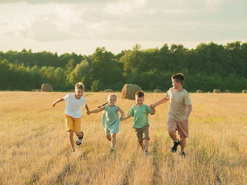 4 Kinder halten sich an den Händen und rennen über ein Feld. Im Hintergrund liegen große Strohballen und man sieht den Wald.